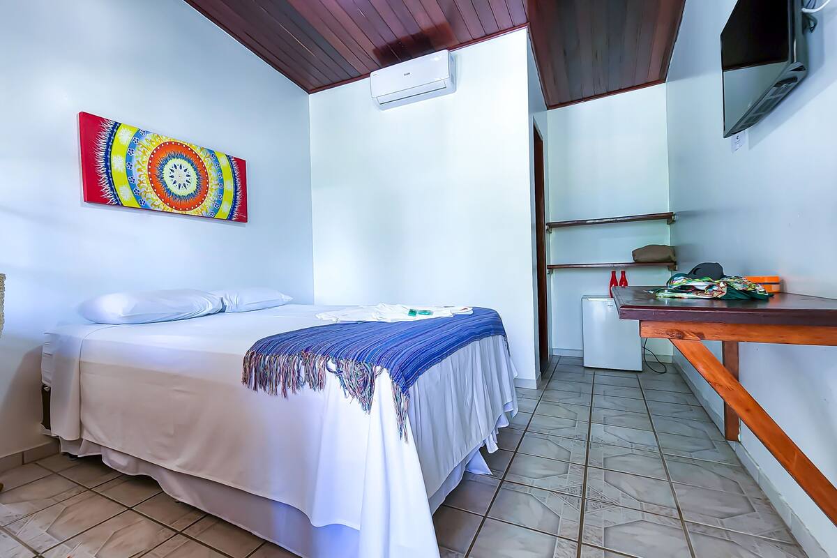 A bedroom with a bed, a colorful artwork on the wall, a small table with items, and a ceiling fan.