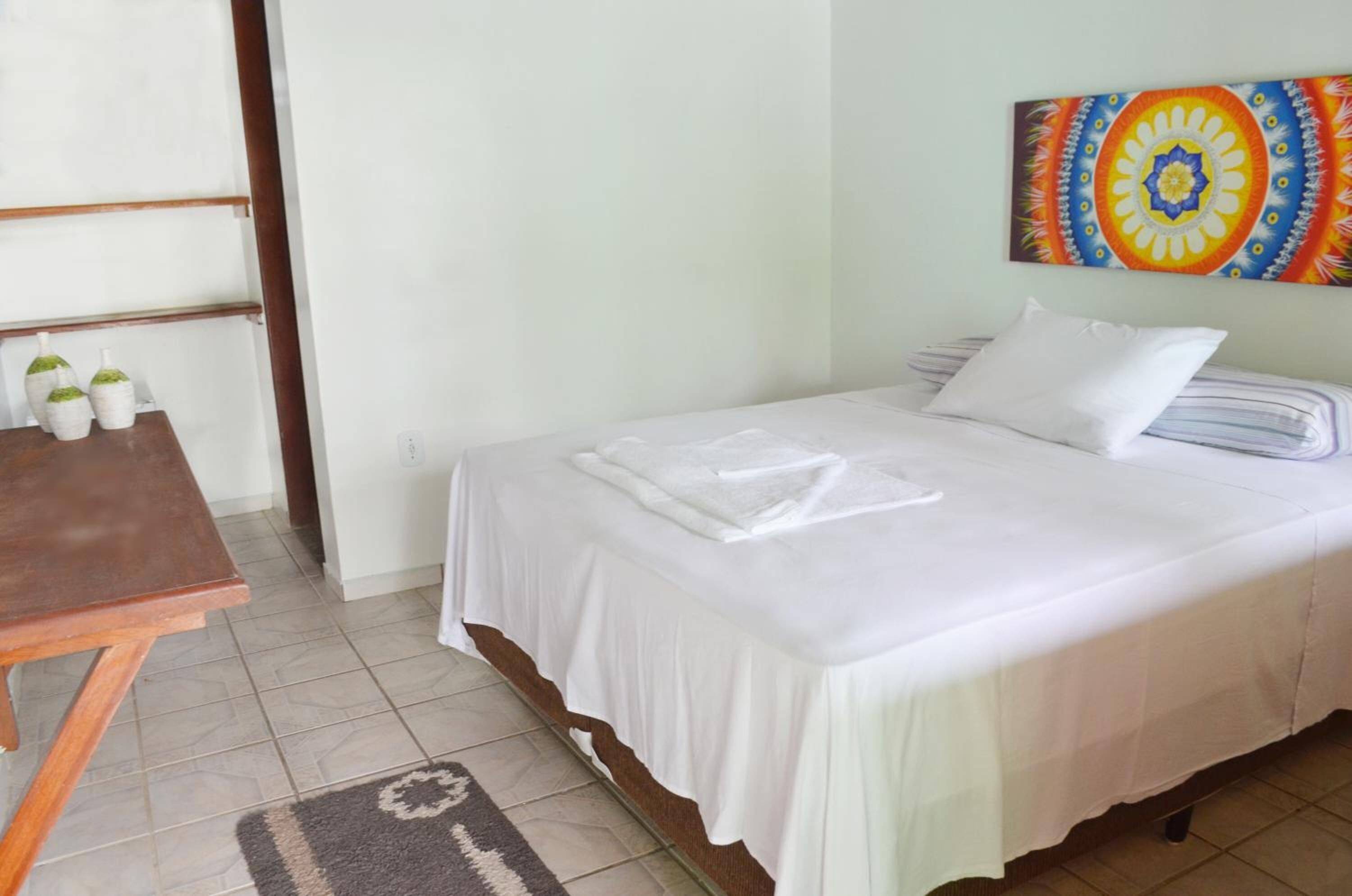 A neatly made bed with white linens, a small table with bottles, and a colorful wall painting.