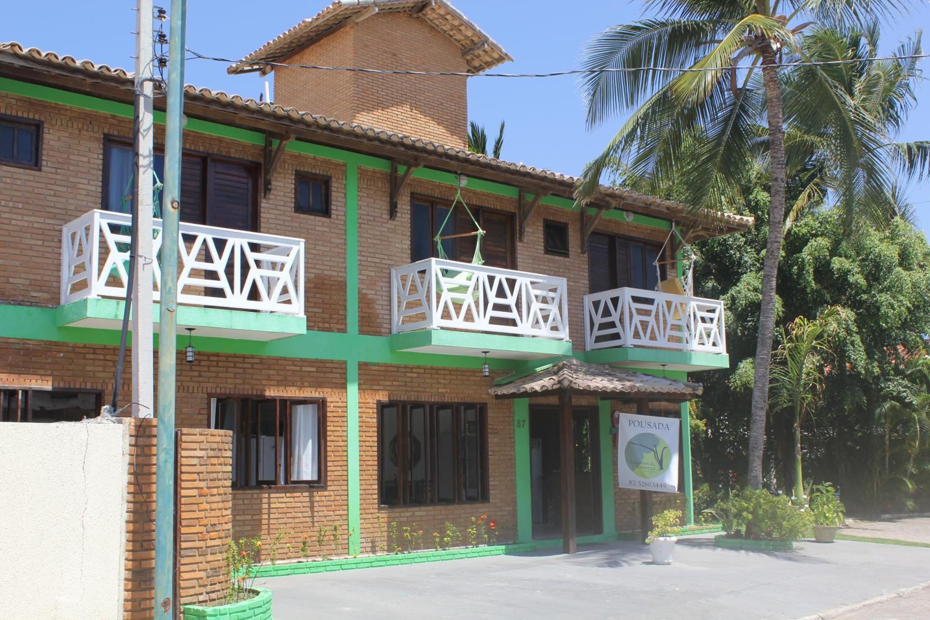 Um edifício de dois andares com fachada verde e marrom, varandas com grades brancas e uma placa com o texto 'POUSADA' e um número de telefone.