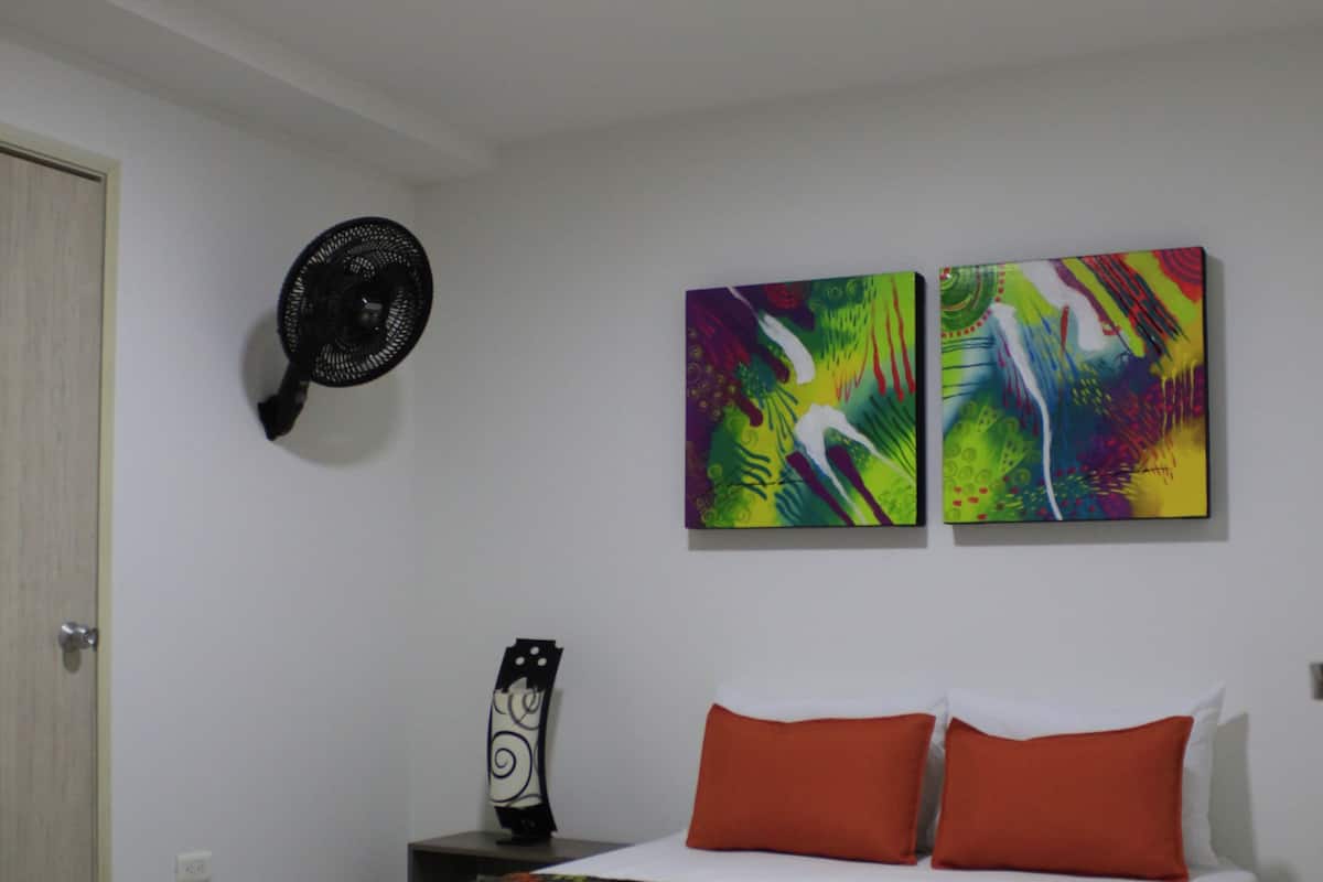 A bedroom with a bed, two wall-mounted artworks, a standing fan, and a nightstand with a decorative item.