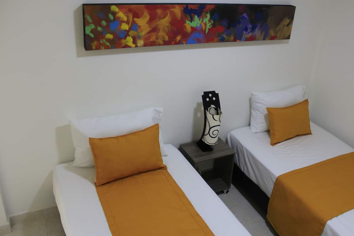 A hotel room with two single beds, each with white bedding and a mustard-colored throw. There is a nightstand between the beds with a decorative item on it. Above the nightstand, there is a colorful abstract painting on the wall.