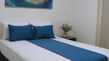 Una cama bien hecha con ropa de cama azul y blanca, una manta de color azul y una pequeña planta en maceta sobre una mesita de noche.