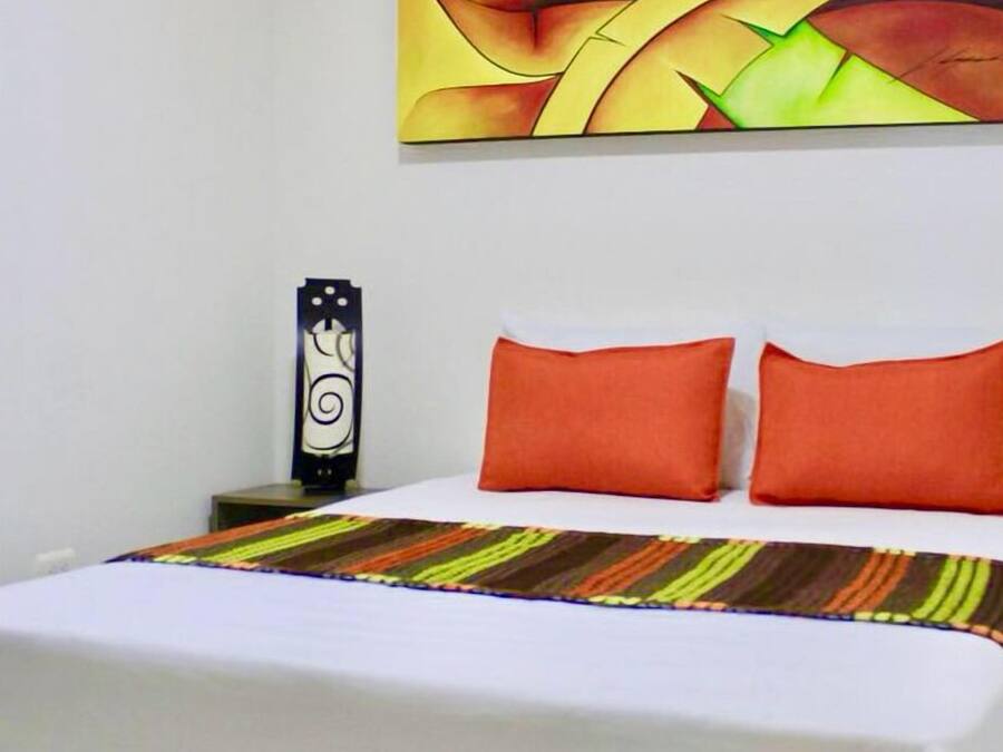 Habitación de hotel con una cama, dos almohadas naranjas, una manta colorida a rayas, una obra de arte colgada en la pared y un objeto decorativo en la mesita de noche.