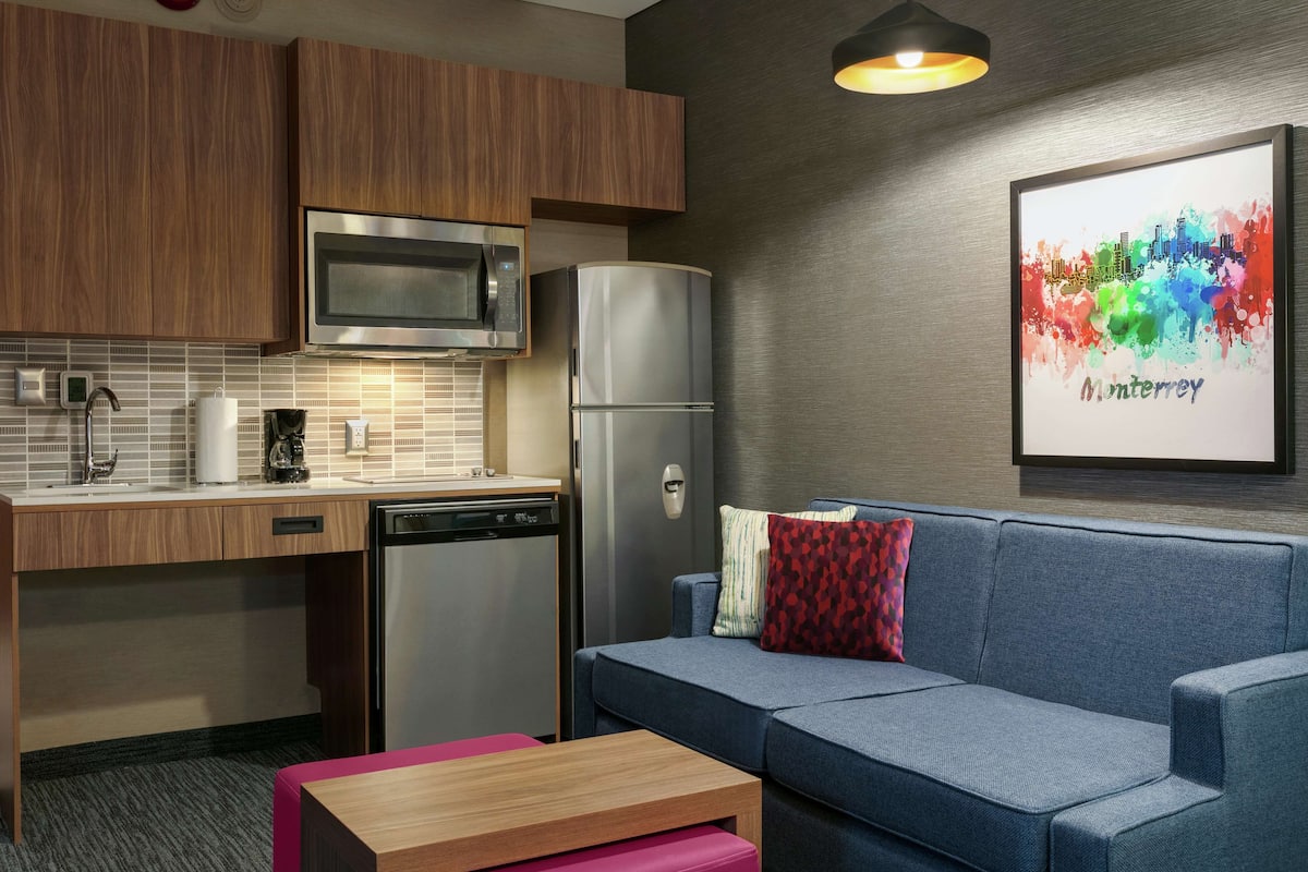 A compact kitchenette with a microwave, refrigerator, and dishwasher. A blue sofa with colorful pillows is against a wall with a framed cityscape artwork.