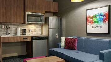 Una kitchenette compacta con microondas, refrigerador y lavavajillas. Un sofá azul con almohadones coloridos está frente a una pared decorada con una obra de arte de un paisaje urbano enmarcado.