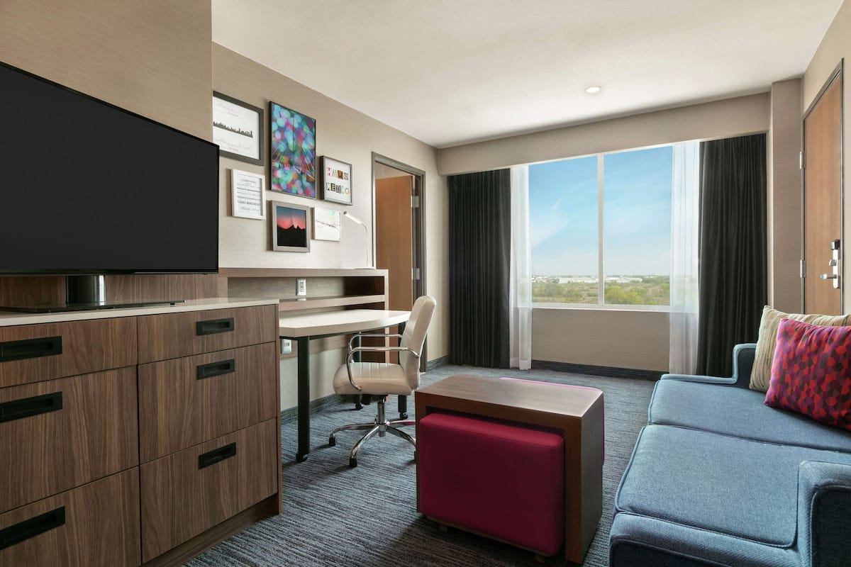A hotel room with a flat-screen TV, a desk with a chair, a sofa, and a large window offering a city view.