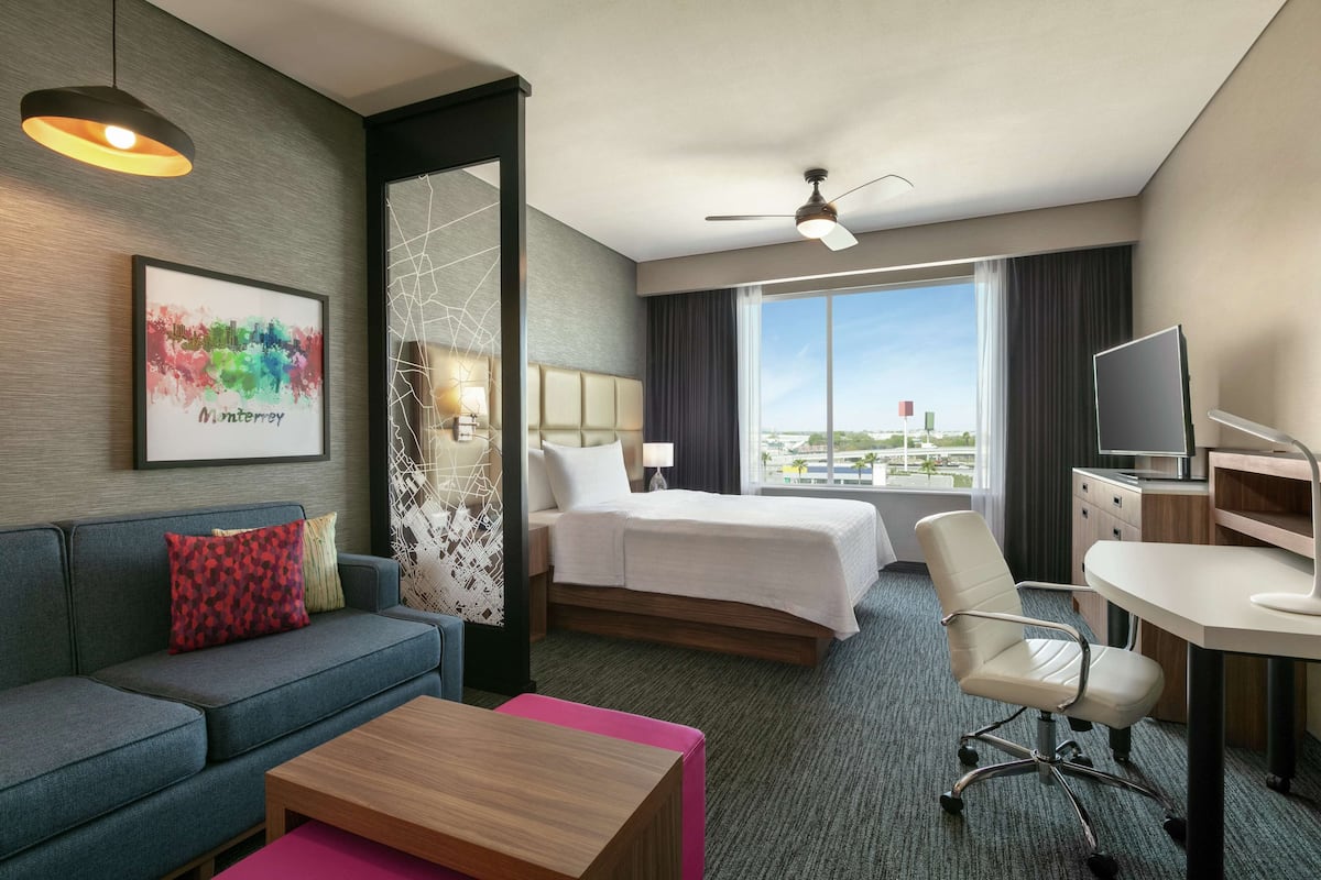 A hotel room with a bed, a sofa, a desk, a chair, a TV, and a large window with a view.