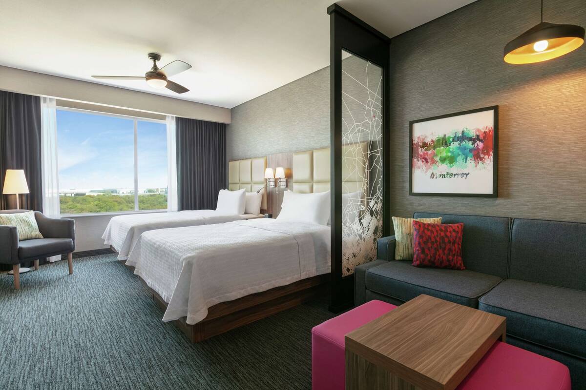 A hotel room with two beds, a sofa, a table, and a large window offering a view.