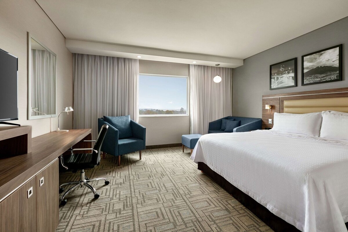Habitación de hotel con cama, escritorio, silla, mesita, espejo y ventana con cortinas.