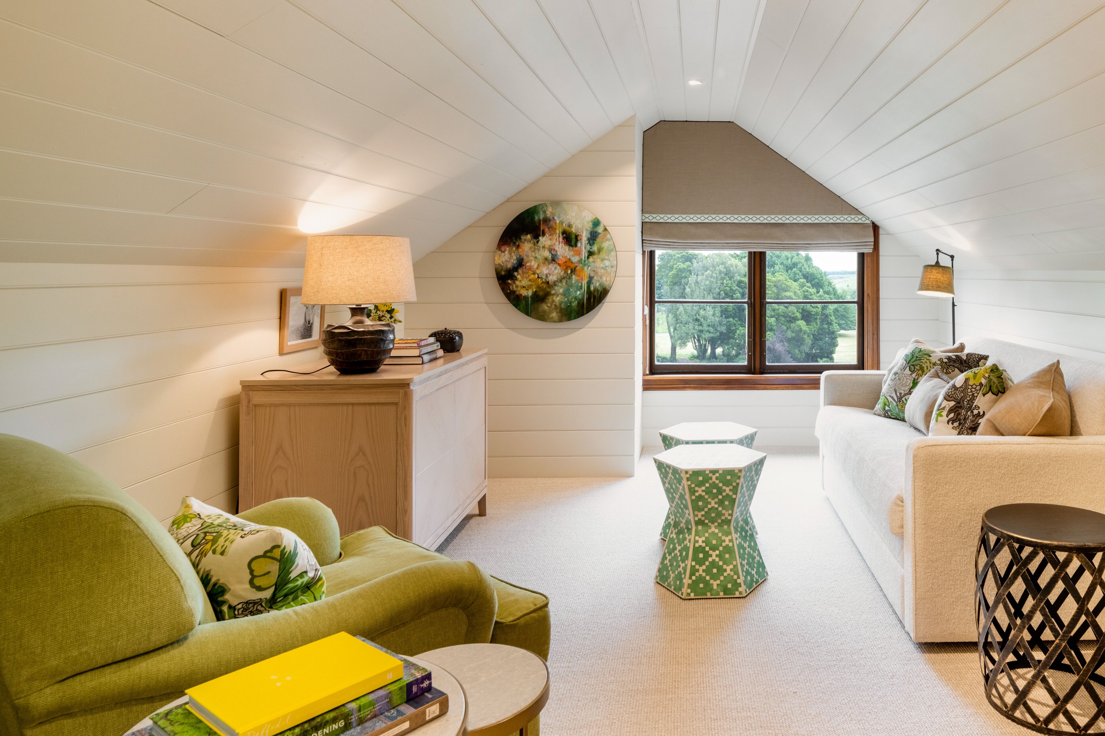 A cozy loft-style room with a high ceiling, a large window with a view of trees, a green sofa, a white couch, and a small round table.