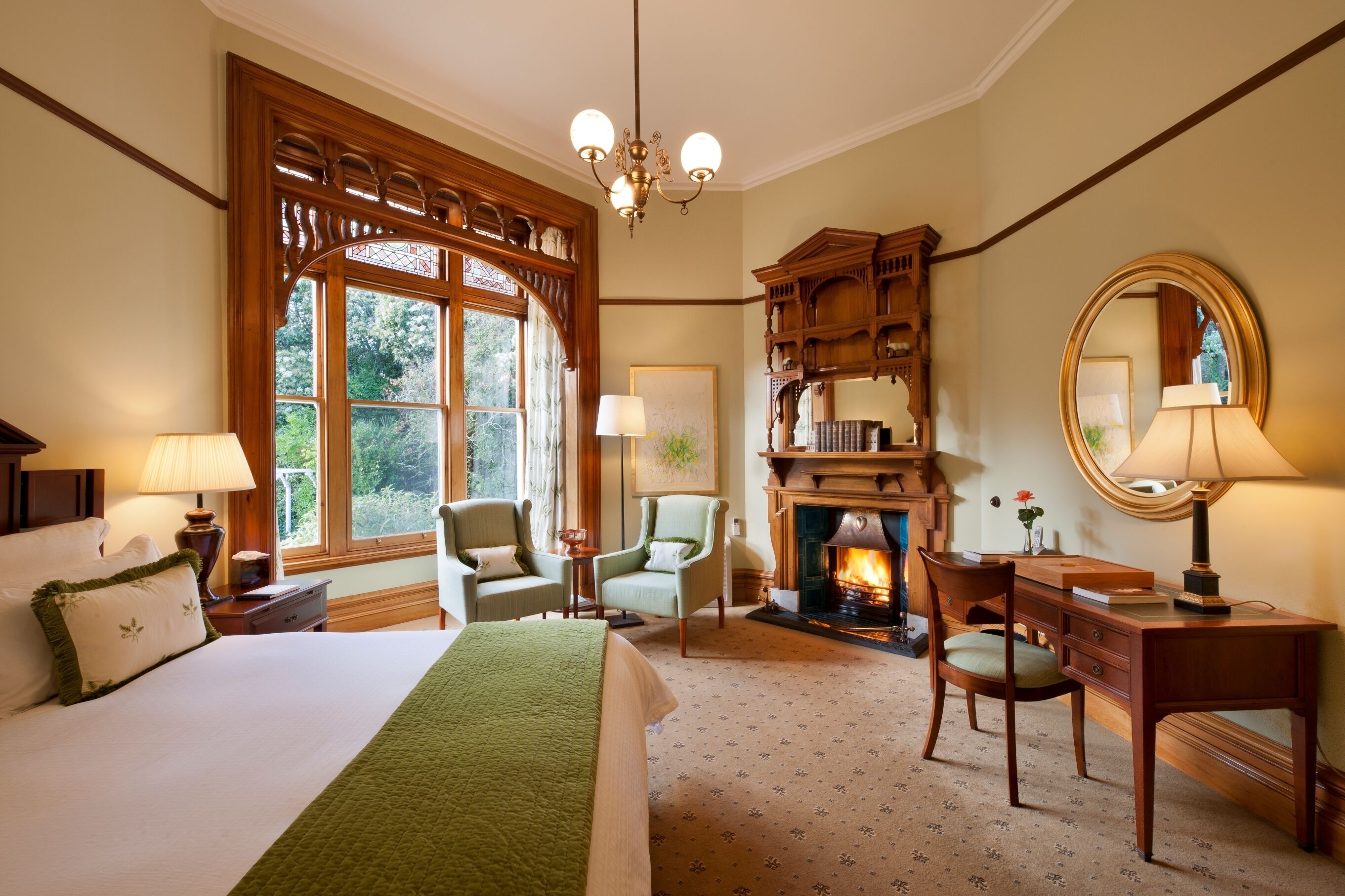 A hotel room with a large bed, two chairs, a desk, a fireplace, and a large window with a view of greenery.
