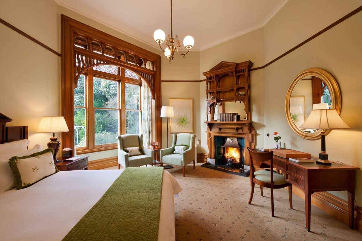A hotel room with a large bed, two chairs, a desk, a fireplace, and a large window with a view of greenery.