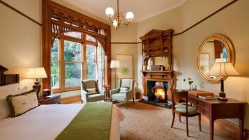 A hotel room with a large bed, two chairs, a desk, a fireplace, and a large window with a view of greenery.