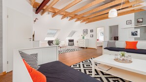 Una habitación moderna y luminosa, con techo alto, vigas de madera, una cama con almohadones estampados, un escritorio y un sofá con un cojín naranja.