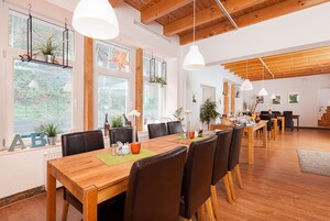 Un comedor amplio con mesa y sillas de madera, ventanas grandes con plantas y lámparas colgantes.