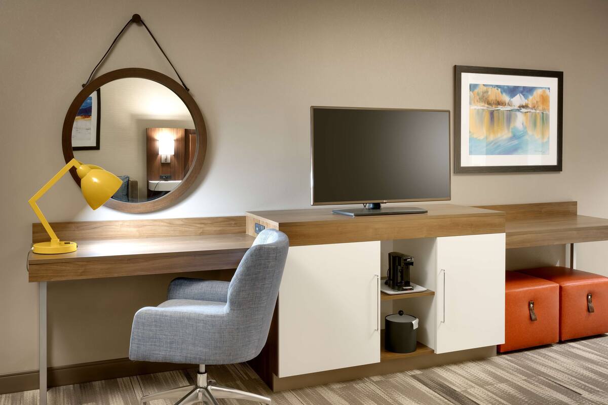 A modern hotel room with a wooden desk, a blue chair, a yellow desk lamp, a flat-screen TV, a framed painting, and storage cabinets.