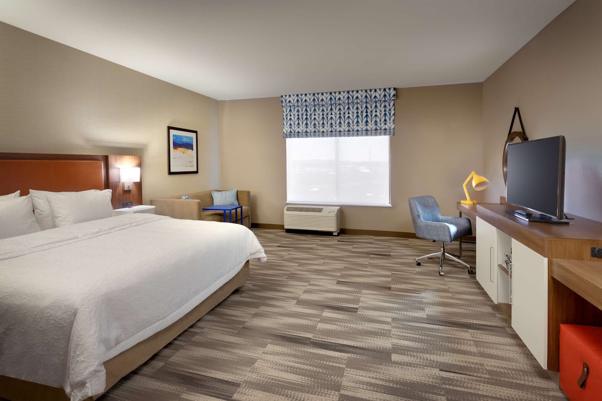 A hotel room with a large bed, a desk with a television, a chair, an air conditioning unit, and a window with a patterned valance.