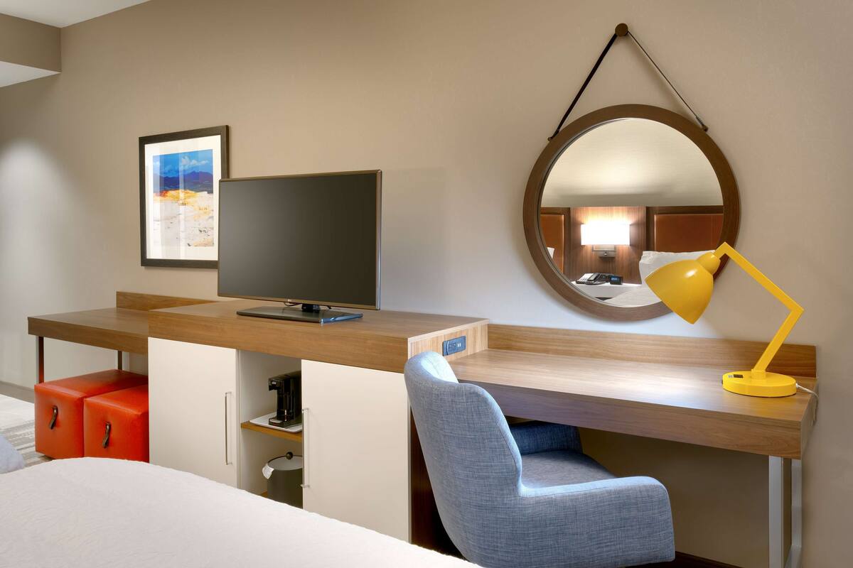 A hotel room with a wooden desk, a television, a mirror, a yellow desk lamp, and a red ottoman.