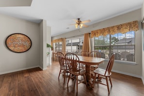 Een eetgedeelte met een houten tafel en stoelen, een plafondventilator en grote ramen met gordijnen.
