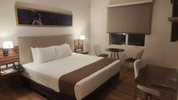 A hotel room with a large bed, bedside tables with lamps, a desk with a chair, a small table, and a ceiling fan.