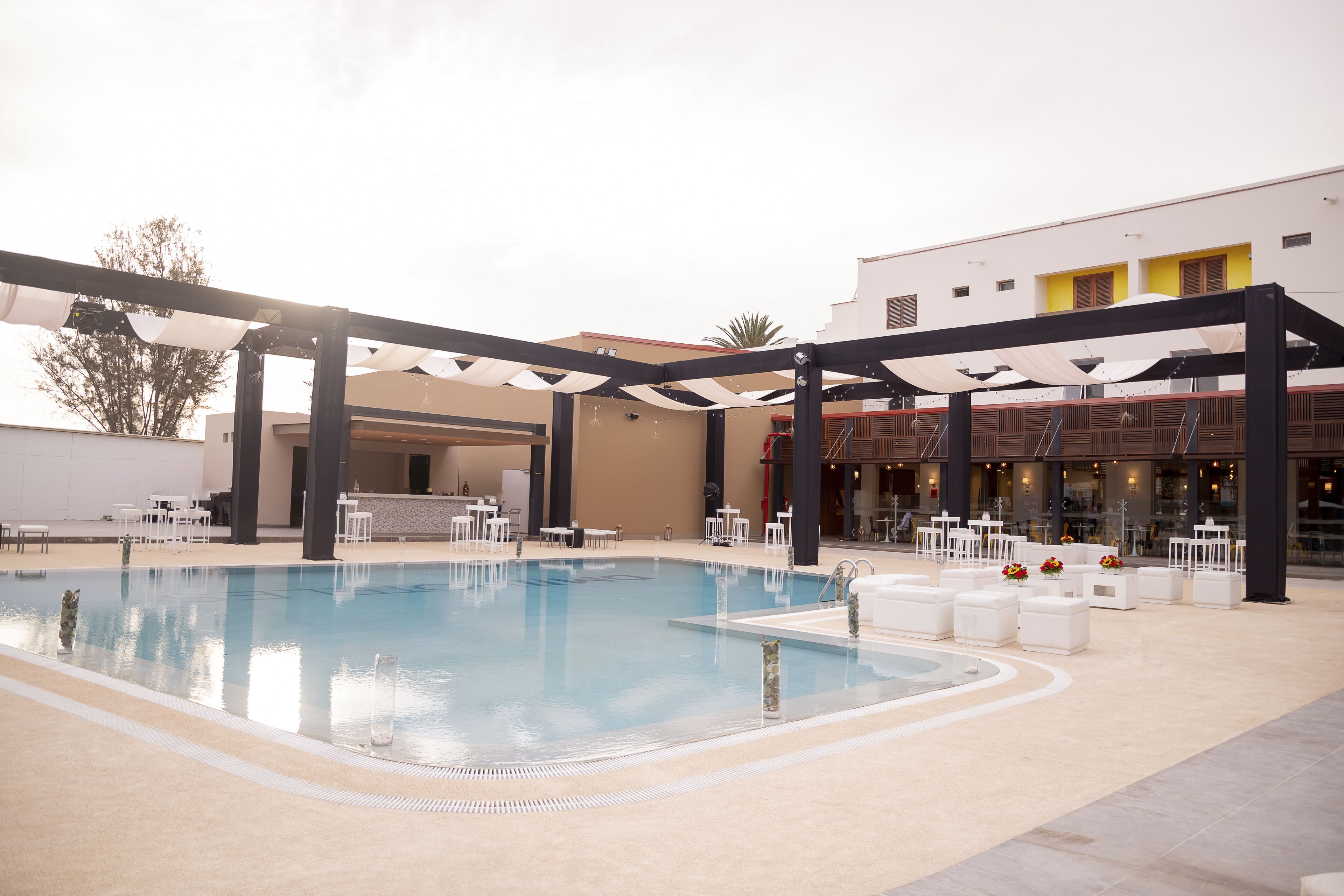 Una piscina con estructura de sombrilla, rodeada de sillones de jardín blancos y mesas, con un edificio de detalles amarillos al fondo.