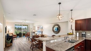 A spacious kitchen with granite countertops, wooden cabinets, and a large island. There is a dining area with a table and chairs, and a view of a landscaped outdoor area through large sliding glass doors. - Fairway S Waikoloa A21 2 Bedroom Condo by RedAwning (Waikoloa)