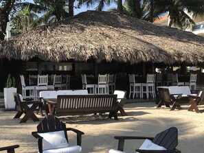Un espacio de descanso frente a la playa con sombrillas de paja, sillas de plástico blanco y bancos de madera.