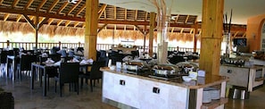 Restaurante em estilo buffet com várias mesas e cadeiras, um balcão central de serviço com recipientes de comida e uma estrutura de telhado de palha.