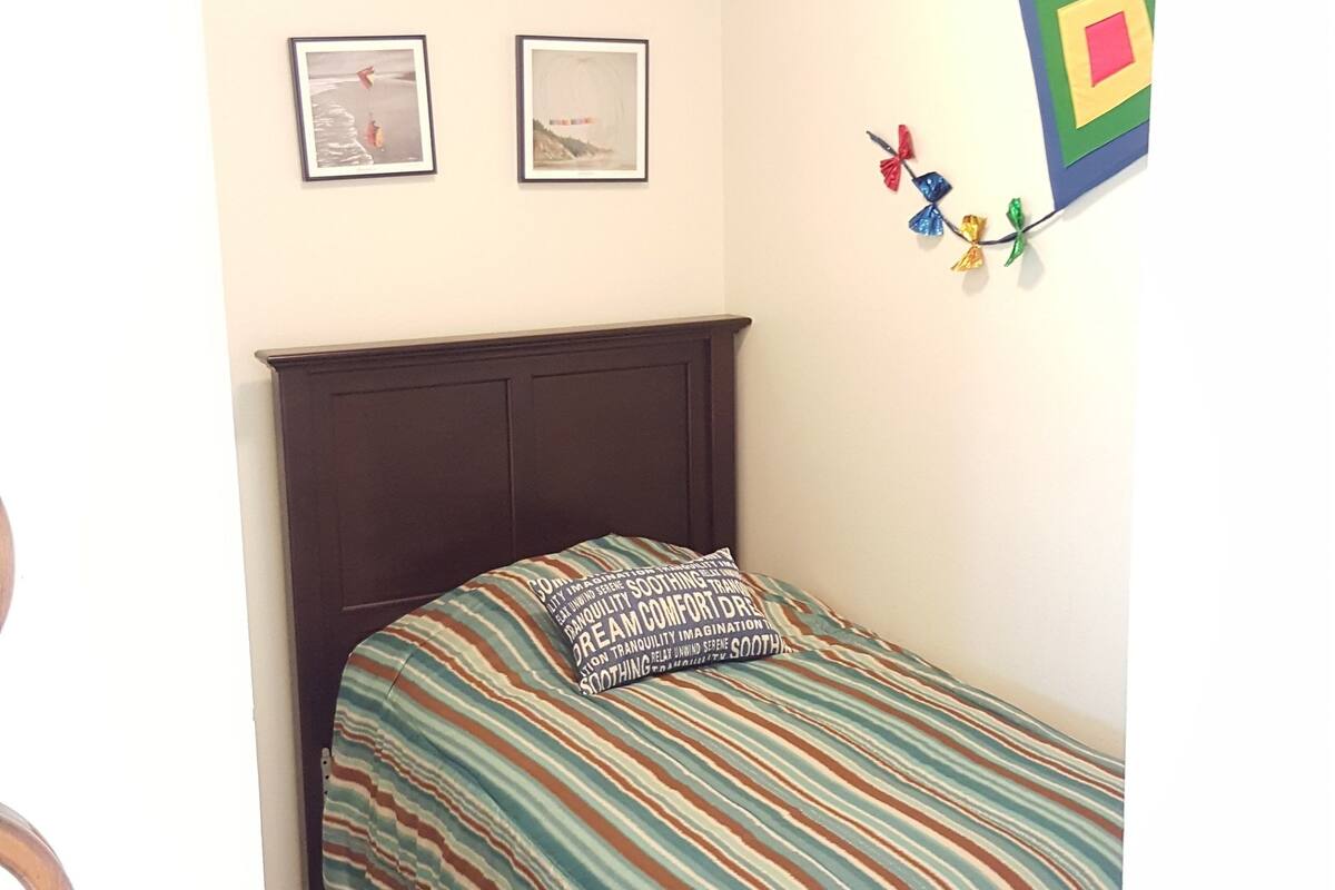 A small, neatly arranged bedroom with a single bed, two framed pictures on the wall, and a ceiling light.
