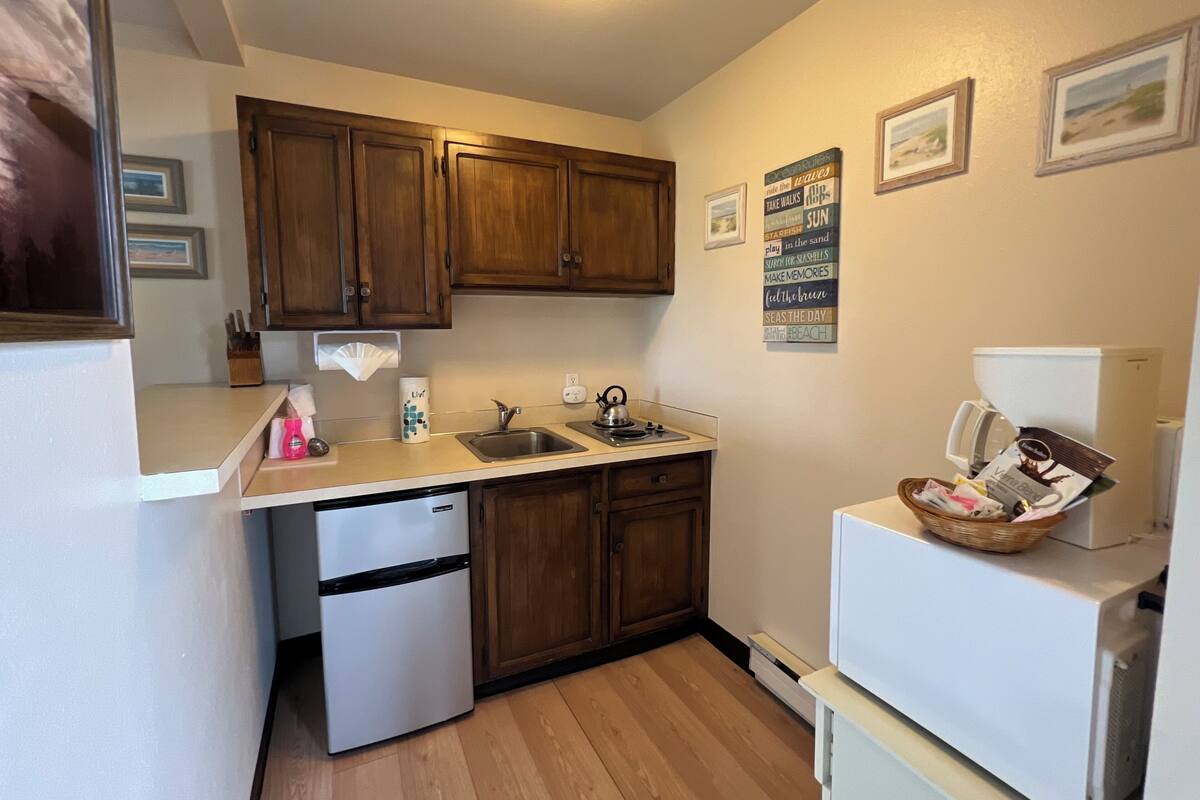A compact kitchen with wooden cabinets, a white refrigerator, a dishwasher, and a sink. There are wall decorations and a basket of items on the counter.