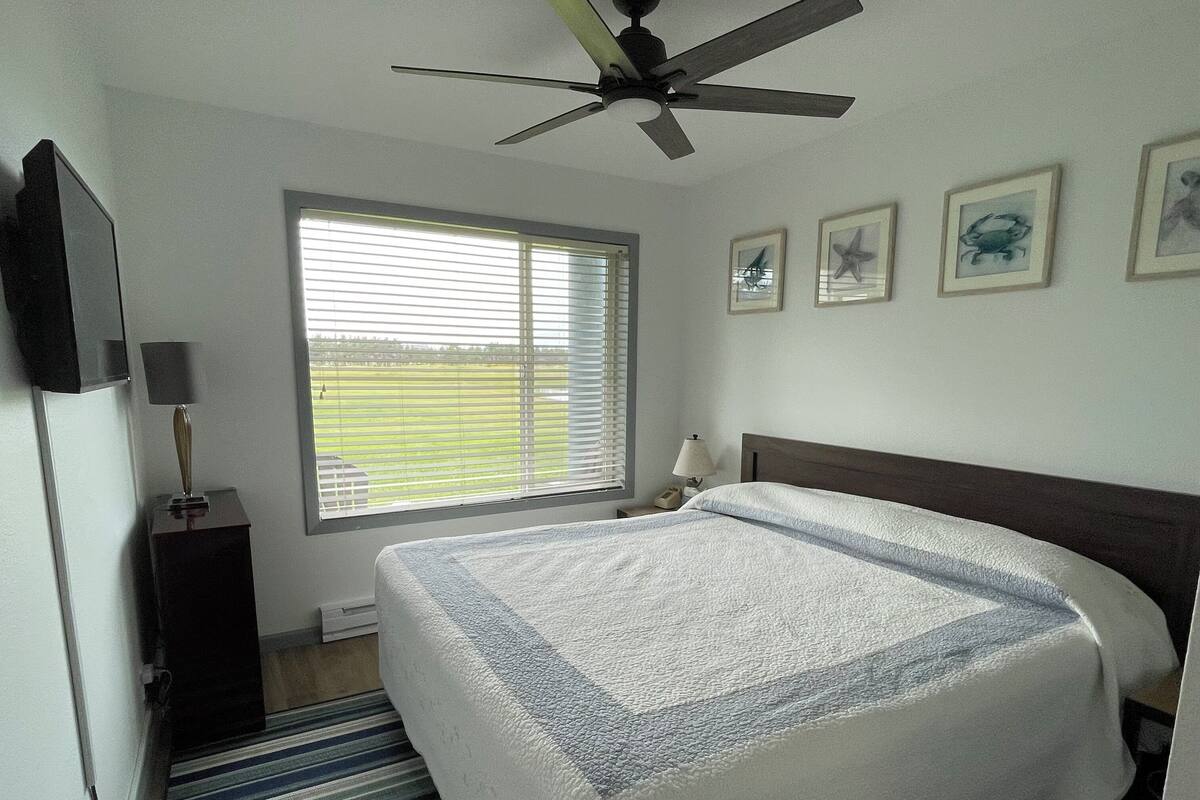 A bedroom with a large bed, a ceiling fan, a window with blinds, and framed pictures on the wall.