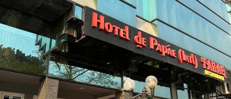 A hotel with a modern glass facade, a sign reading "Hotel de Papas (And) Tabao Restaurant", and outdoor seating with red chairs.