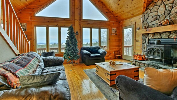 Interior acogedor de una cabaña con chimenea de piedra, un árbol de Navidad y un ventanal con vistas a un paisaje nevado.