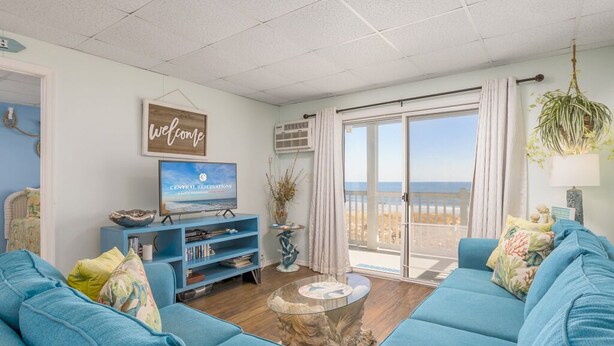 A living room with a blue sofa, a television, and a view of the beach through a sliding glass door. - Oceanfront condos with 3 night minimum rentals and linens included! (Ocean City)