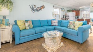 A living room with a blue sectional sofa, a glass coffee table, and a wall mural. - Oceanfront condos with 3 night minimum rentals and linens included! (Ocean City)