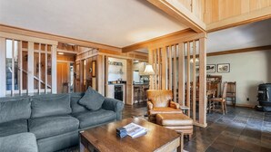 Un vaste salon avec un canapé gris, une table basse en bois et un fauteuil en cuir. Un escalier mène à l’étage supérieur.