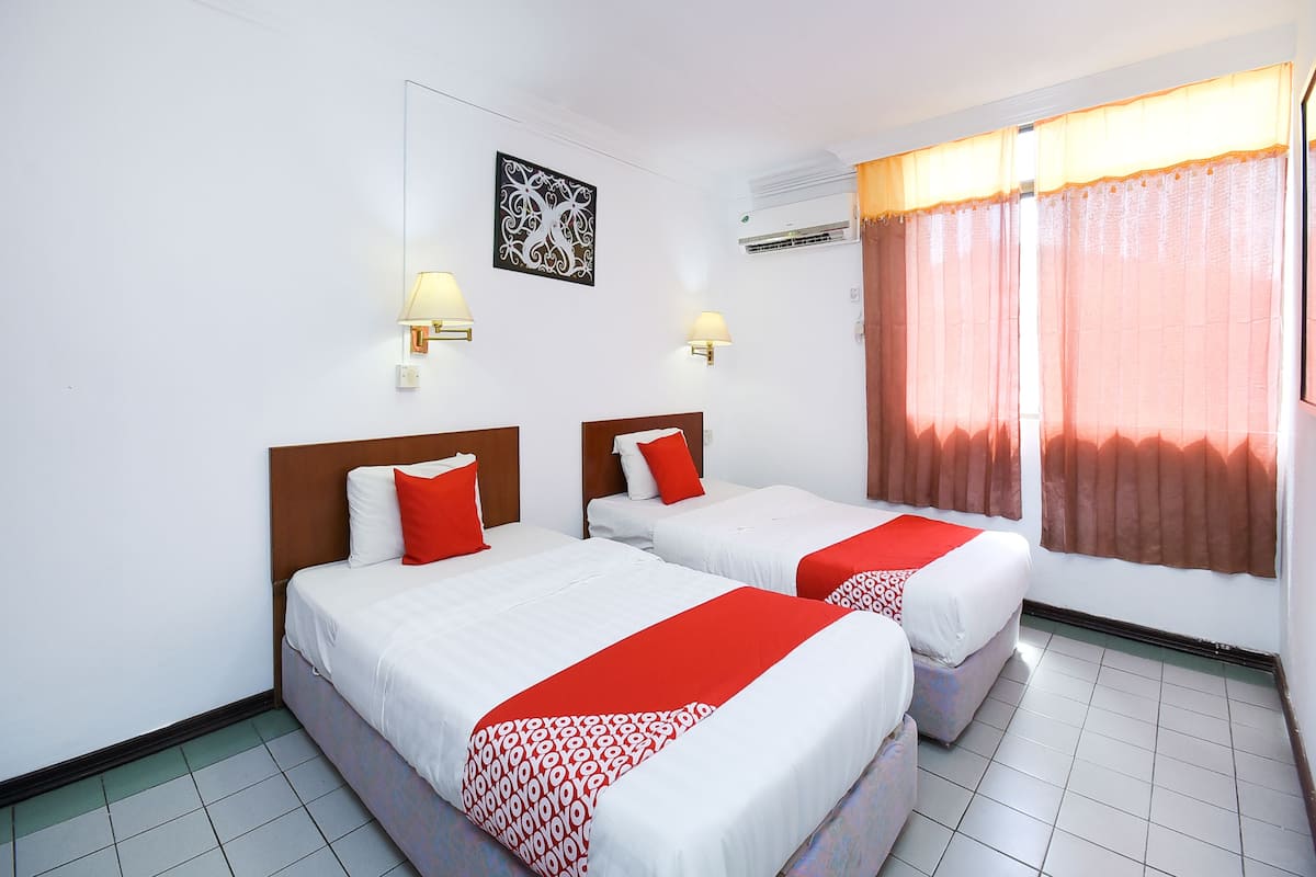 Une chambre d’hôtel avec deux lits, du linge de lit blanc, des touches de couleur rouge, des lampes fixées au mur, un appareil de climatisation et une fenêtre avec des rideaux.