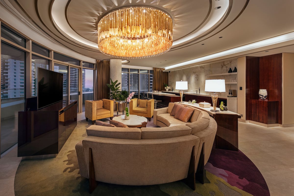 A modern living room with a large TV, comfortable seating, a chandelier, and a view of the city.