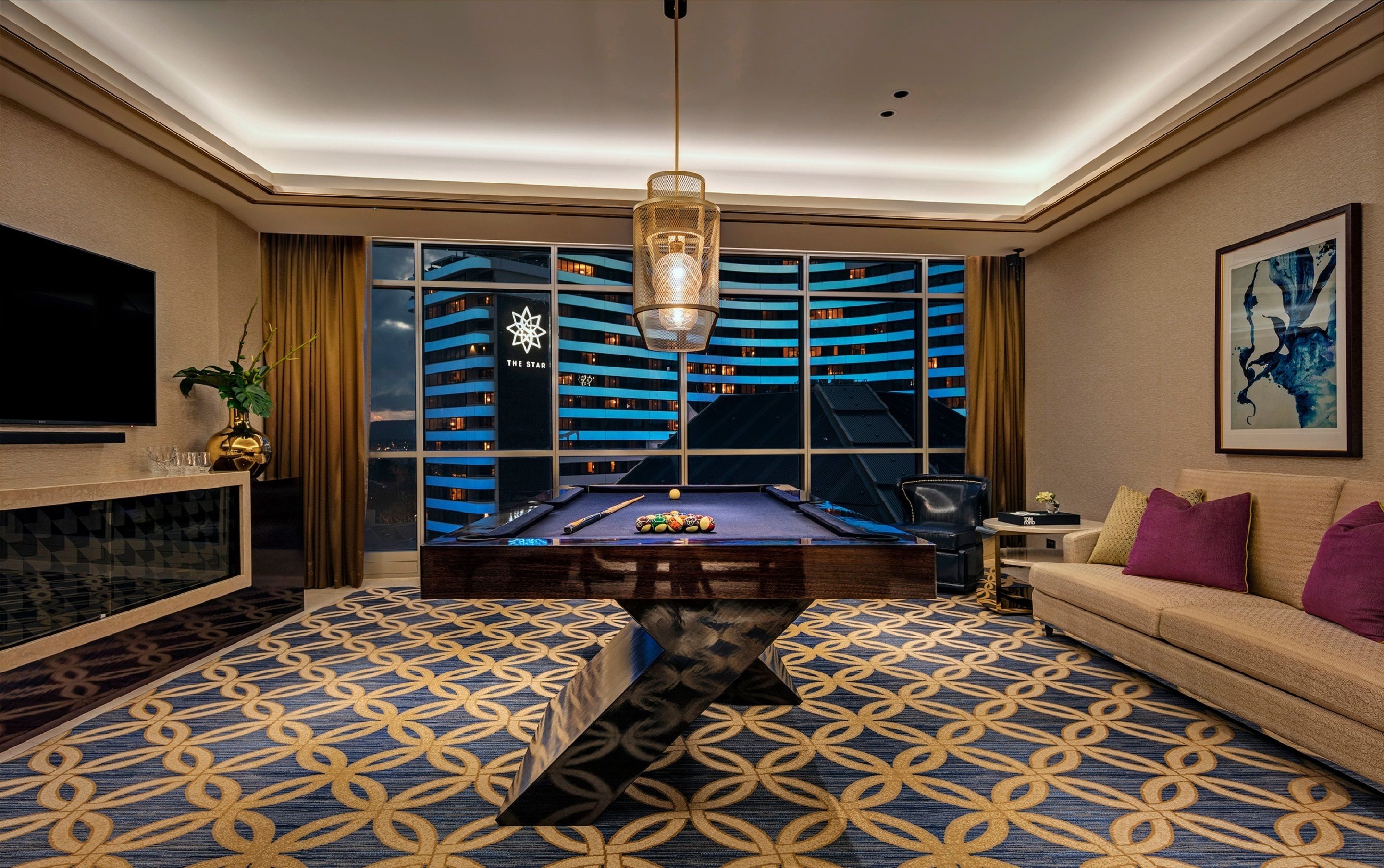 A modern hotel room with a pool table, a sofa, a TV, and a large window overlooking a cityscape.