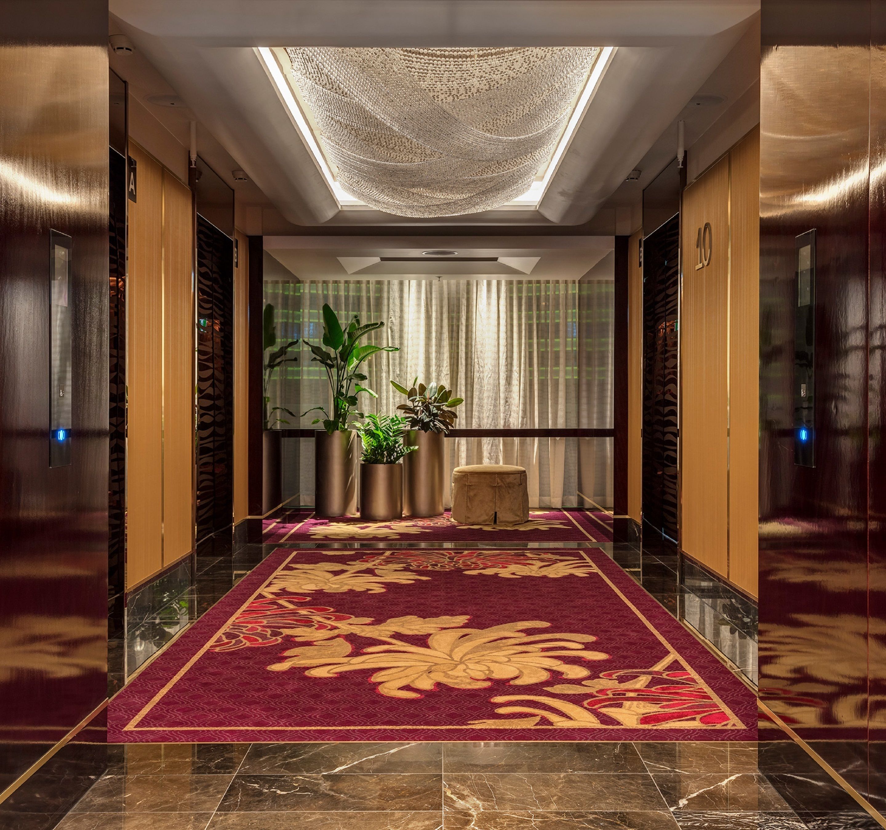 A hotel lobby with a marble floor, a red carpet with gold patterns, potted plants, and a large chandelier.