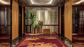 A hotel lobby with a marble floor, a red carpet with gold patterns, potted plants, and a large chandelier.