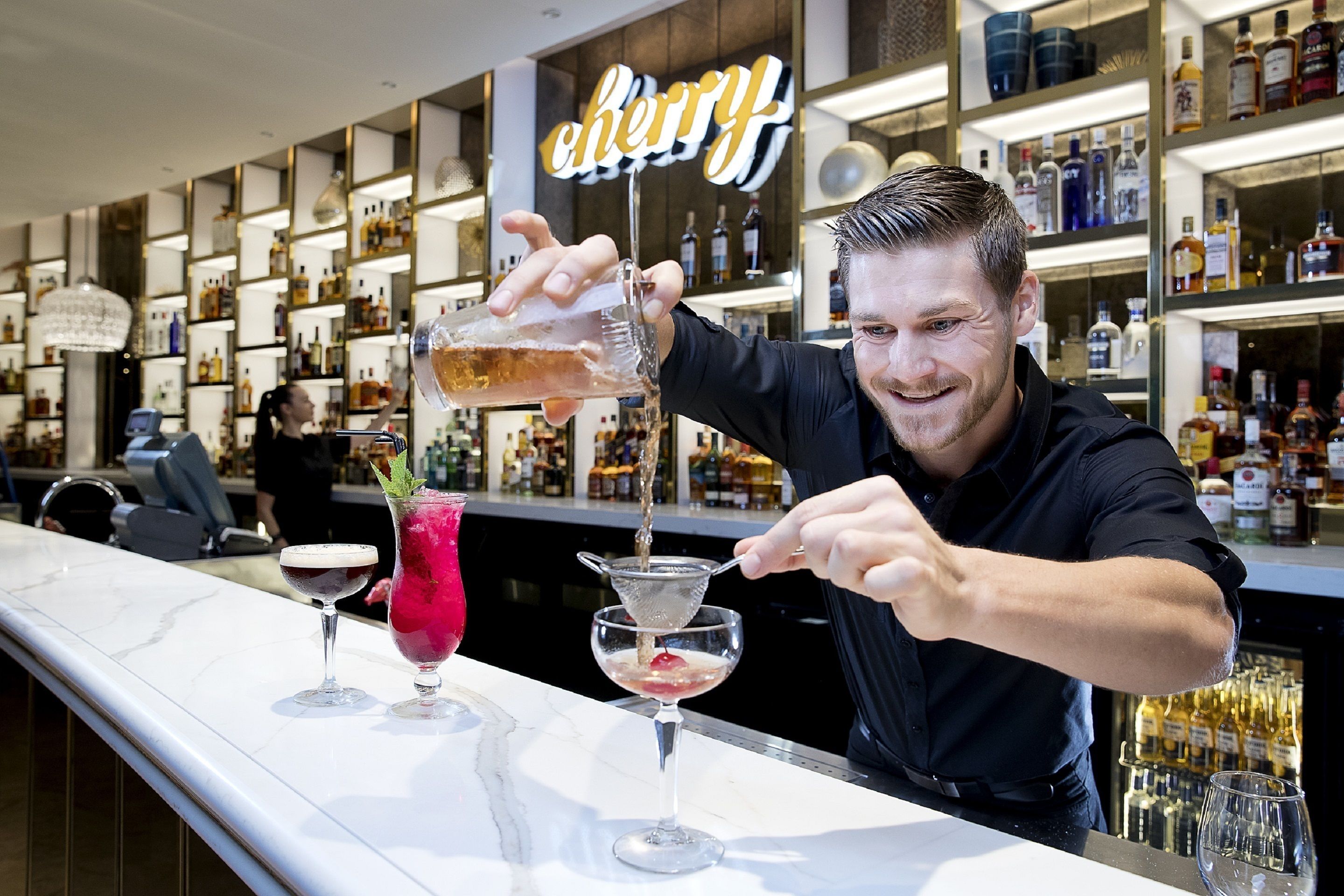 Un barman verse une boisson dans un verre au comptoir d’un bar, avec des étagères remplies de bouteilles d’alcool en arrière-plan.