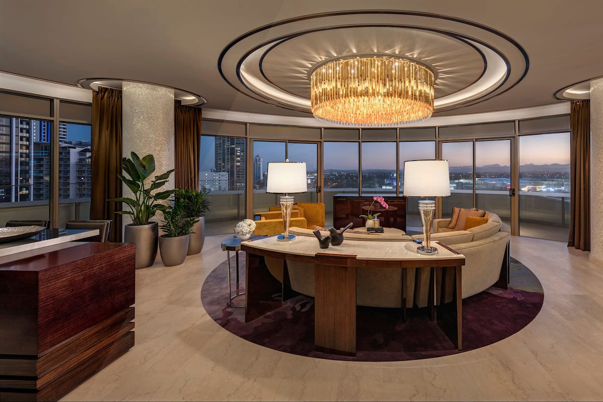 A modern hotel room with a round dining table, two lamps, a chandelier, a sofa, and a view of the cityscape.