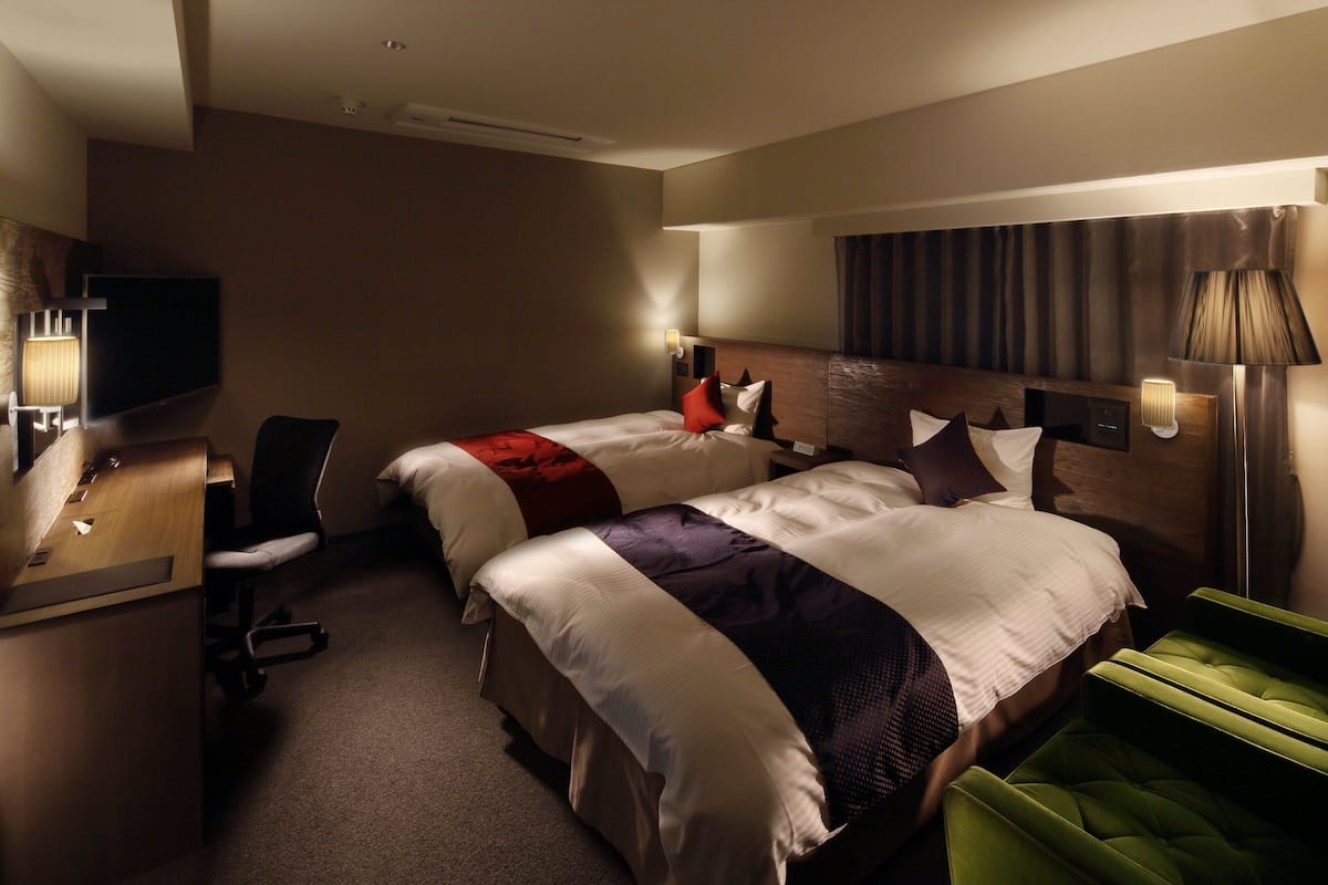 Quarto de hotel com duas camas, uma mesa, uma cadeira e uma televisão.