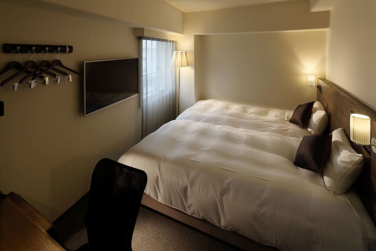 Quarto de hotel com uma cama grande, uma televisão fixada na parede, um cabideiro com ganchos e uma janela com cortinas.
