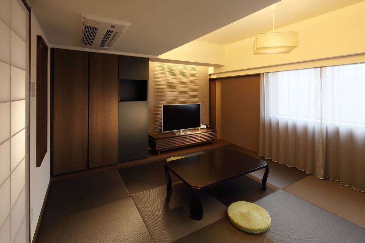 Quarto moderno e compacto com TV de tela plana, uma mesa pequena e uma cama tipo futon.