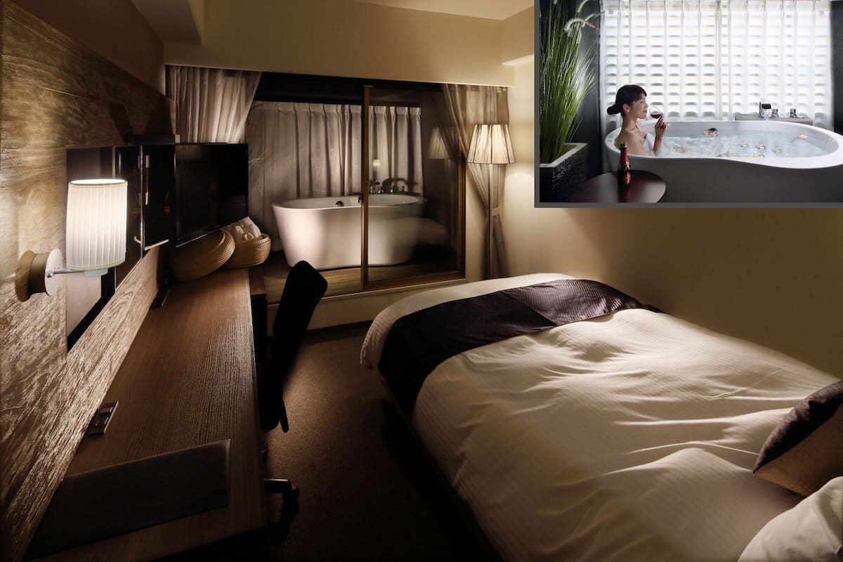 Quarto de hotel moderno com uma cama grande, banheiro separado com banheira, televisão de tela plana e uma janela ampla com cortinas.