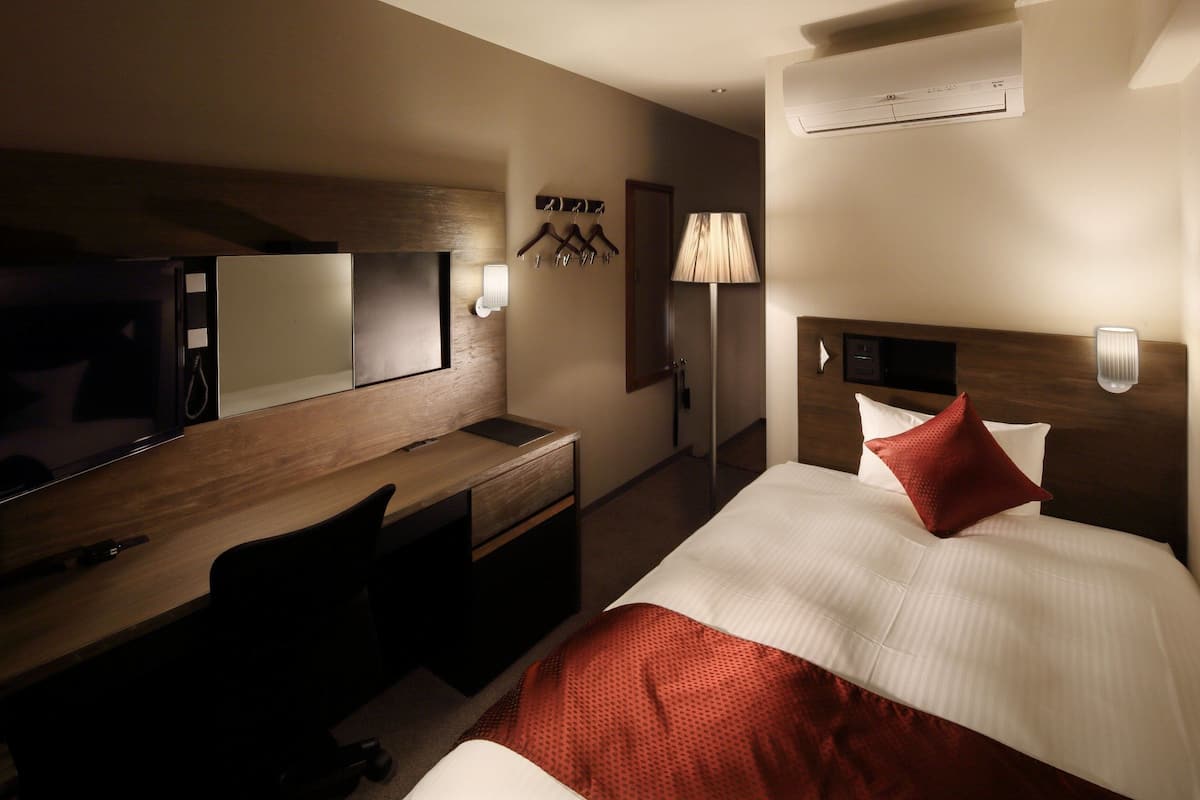 Quarto de hotel com cama, escrivaninha, cadeira, espelho, luminária e ar condicionado fixado na parede.