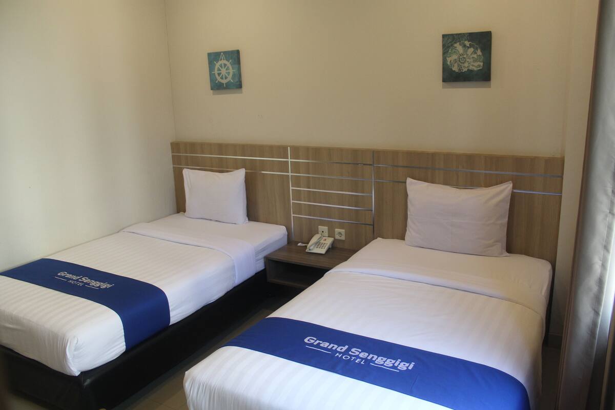 A hotel room with two single beds, each with a blue bedspread and white pillows. There is a wooden headboard, a bedside table with a phone, and a wall-mounted artwork.