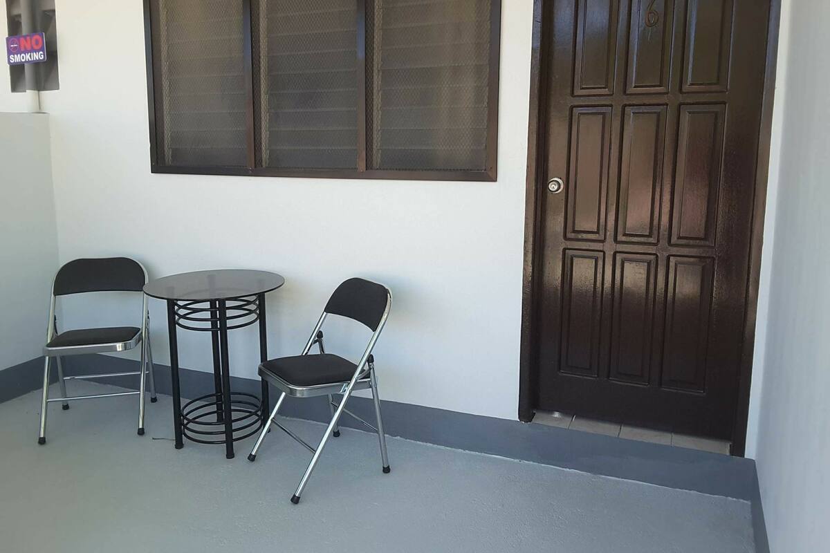 Una habitación con una puerta de madera oscura, una ventana con reja de metal y dos sillas junto a una pequeña mesa.